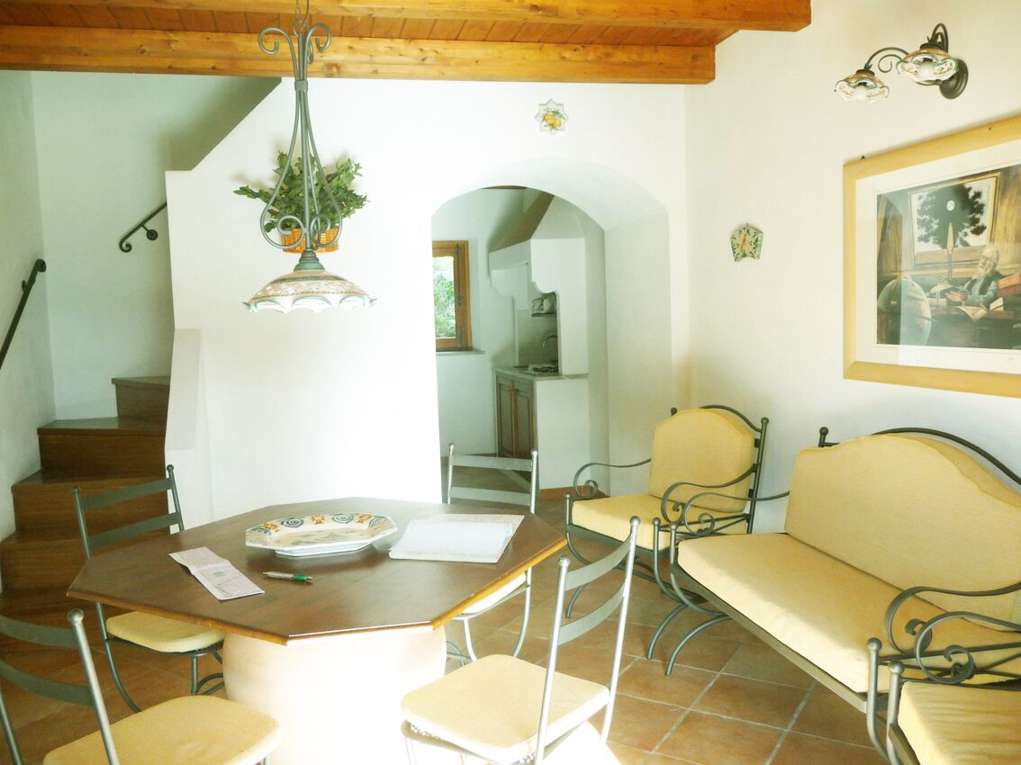 Zona pranzo con tavolo in legno, sedie e divano. Una scala conduce a un'altra stanza, con un lampadario sopra il tavolo.