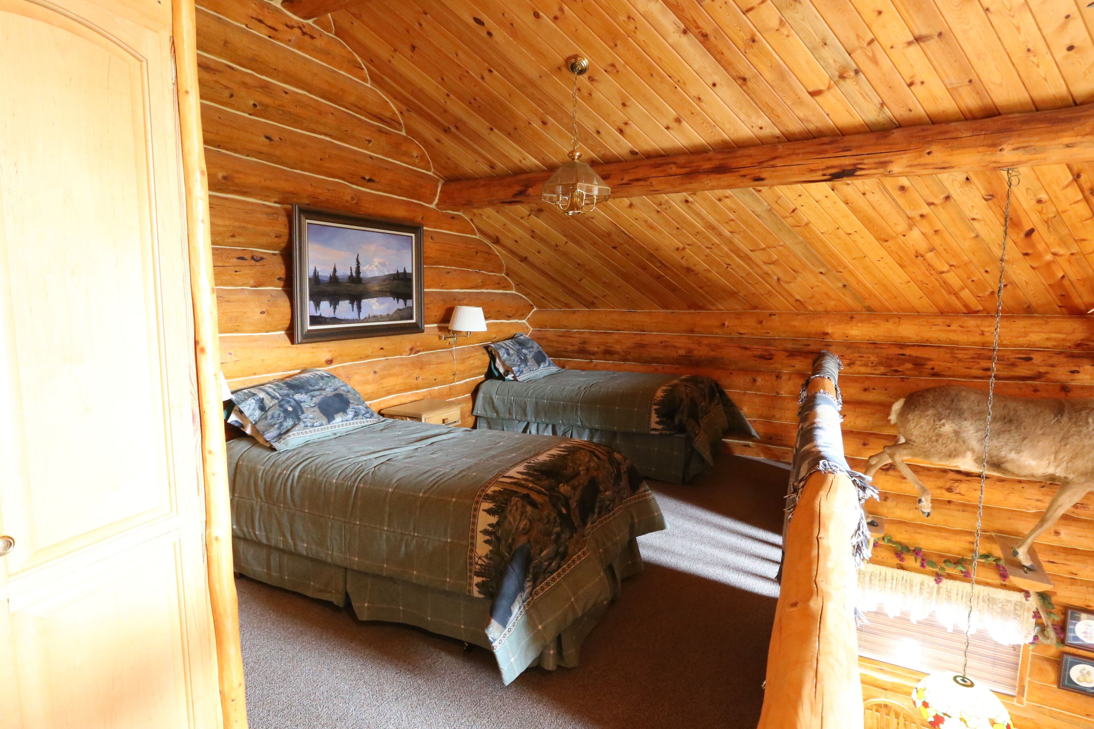 A log cabin bedroom with two beds, a hanging light fixture, and a deer in the ceiling.