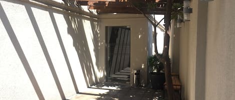 Een smalle buitentuin met een houten pergola erboven, een plant in pot en een prullenbak.