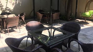 Un patio con mobiliario de mimbre, incluyendo una mesa y sillas con superficie de vidrio, rodeado por un muro bajo y una pérgola cubierta de plantas trepadoras.