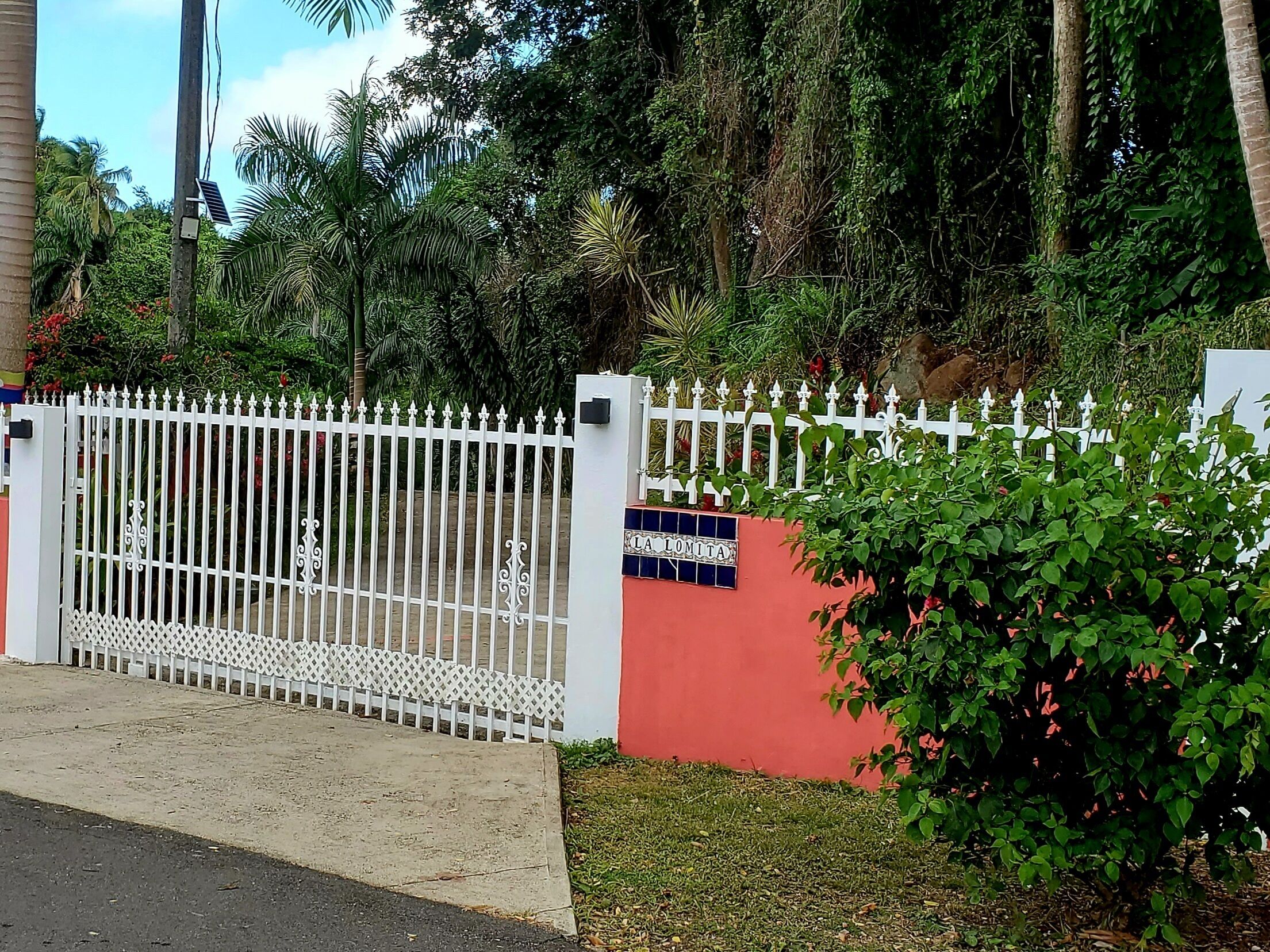 A gated entrance with a white picket fence, a red wall, and lush greenery.