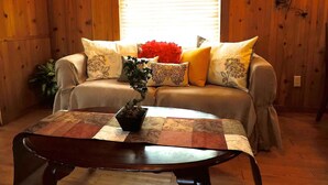 A cozy living room with a wooden floor, a sofa with patterned cushions, a coffee table with a plant, and a window with blinds. - Wildflowers Cabin @ HomeAway Ranch (Boerne)