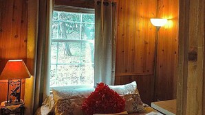 A cozy wooden cabin bedroom with a bed, a window with curtains, a bedside lamp, and a ceiling light. - Wildflowers Cabin @ HomeAway Ranch (Boerne)