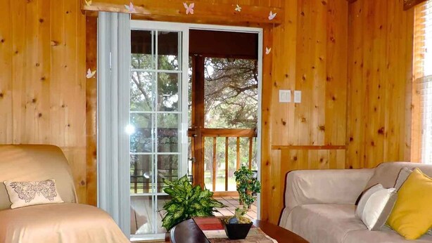 A cozy cabin interior with wooden walls, a wooden ceiling, a large window, a sofa, a bench, and a table with a plant. - Wildflowers Cabin @ HomeAway Ranch (Boerne)