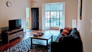 Un salon comprenant un téléviseur, un canapé, une table basse, un tapis à motifs, un ventilateur de plafond et une grande fenêtre avec des stores.