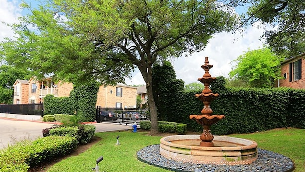 Une fontaine à plusieurs niveaux, entourée d’une bordure circulaire en pierre, est installée dans un espace verdoyant bien entretenu, agrémenté de haies taillées et d’un grand arbre en arrière-plan.