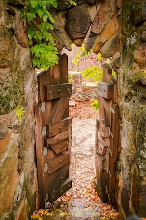Un arc en pierre surmonté de portails en bois, qui mène à un sentier recouvert de feuilles mortes.