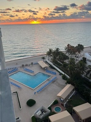 Un espace piscine d’un complexe hôtelier offrant une vue sur le coucher de soleil sur l’océan, avec des palmiers et des chaises longues.