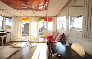A modern living room with a sofa, dining table, and chairs. There is a large window with curtains, and a view of buildings outside.