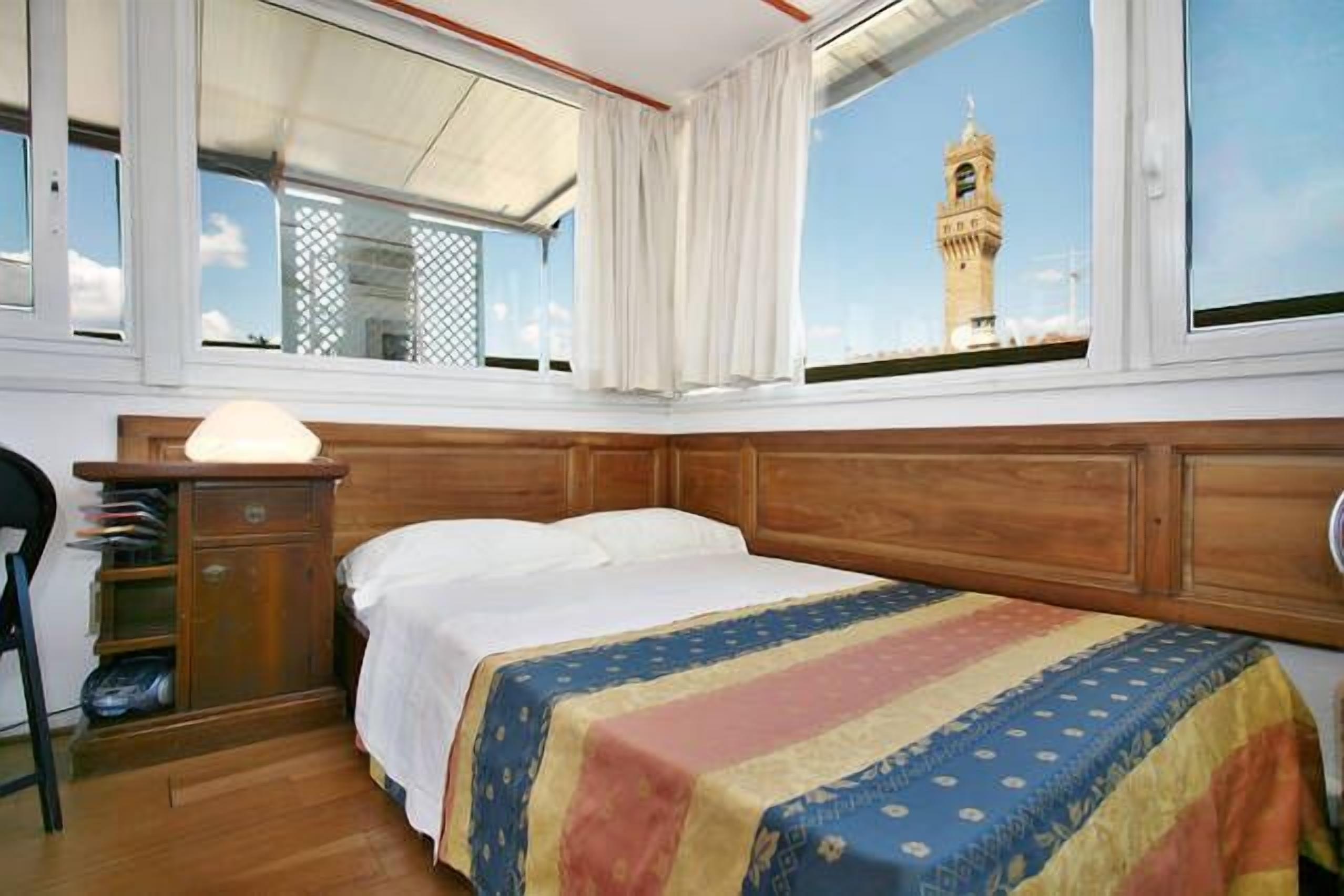 A bedroom with a large window, a wooden headboard, a bed with a colorful bedspread, and a nightstand with a lamp.
