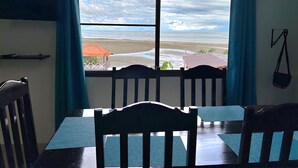 A room with a large window overlooking a beach. There is a table with chairs in front of the window.