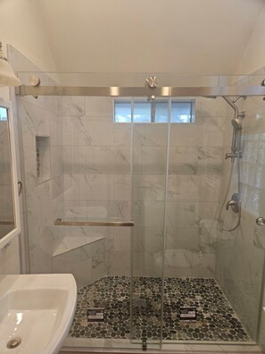 A glass-enclosed shower with a handheld showerhead, a small window, and a tiled floor with a pebble pattern.