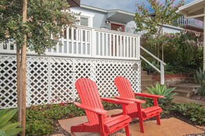 Una casa con cerca blanca, dos sillas Adirondack rojas y un jardín con diversas plantas.