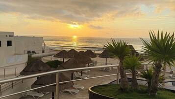 A balcony with a view of the sunset over the sea, a large planter with palm plants, and thatched umbrellas.