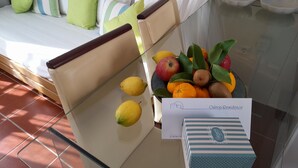 A hotel room with a bed, pillows, a glass table with fruits and a card, and a bedside table with a bowl.