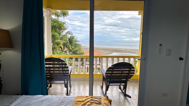 A room with a bed, a balcony with two chairs, and a view of the beach.