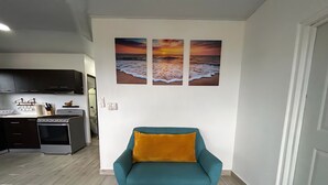 A modern living room with a teal sofa, a kitchen in the background, and a sunset painting on the wall.