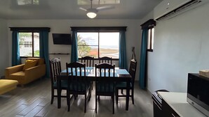 A dining area with a table and chairs, a sofa, a TV, and a view of the beach.