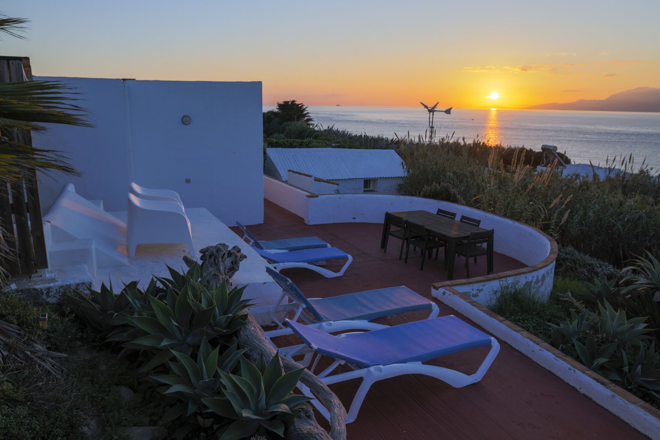 An outdoor seating area with lounge chairs and a table, overlooking a body of water at sunset.