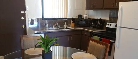 A compact kitchen with a sink, microwave, stove, and refrigerator. There is a dining table with chairs and a potted plant.