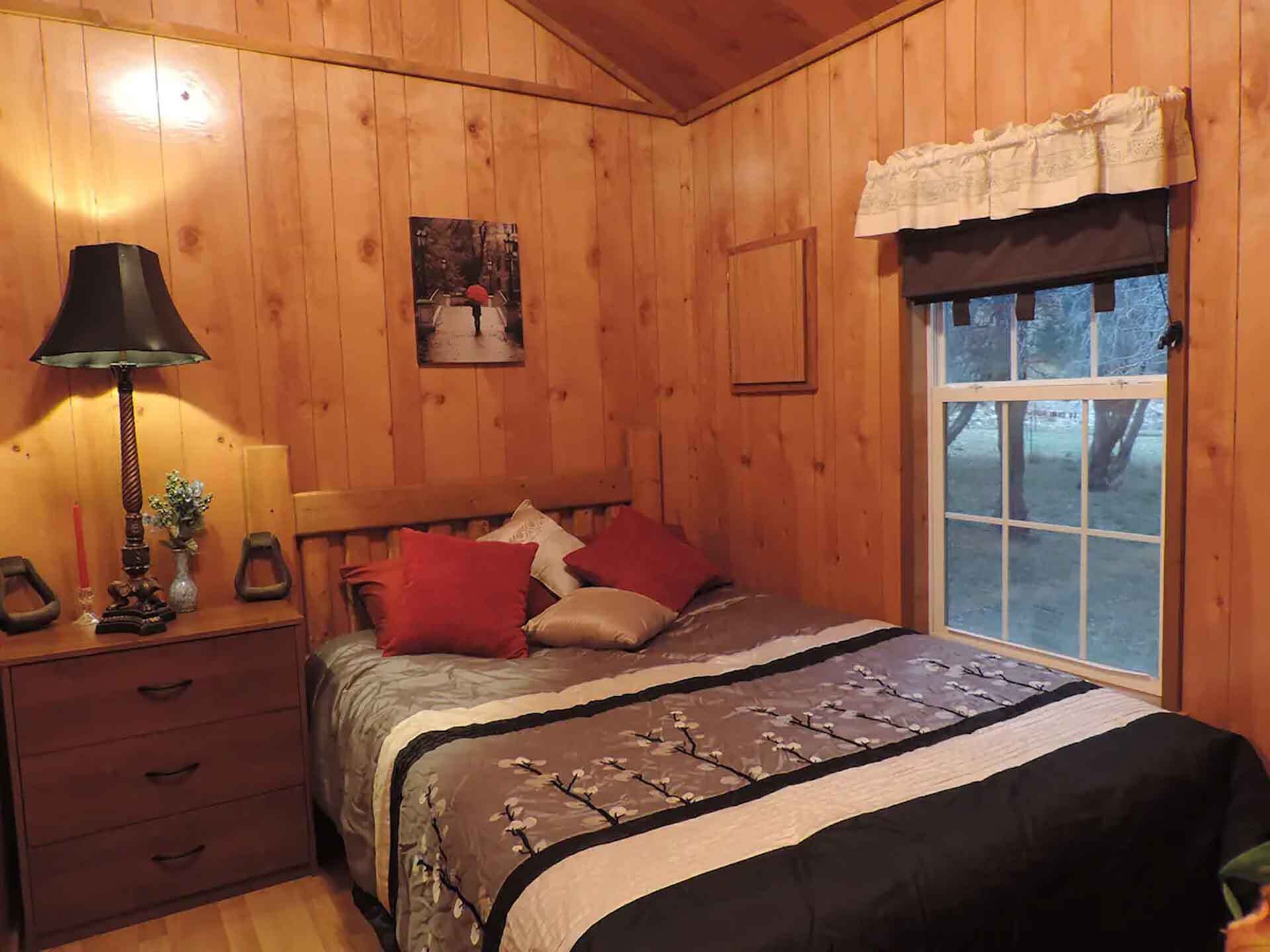 Une pièce en bois avec un lit, une table de chevet, une lampe, une fenêtre et un tableau accroché au mur.
