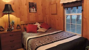 A wooden room with a bed, a nightstand, a lamp, a window, and a painting on the wall. - Hansel & Gretel Charming Cabin @ HomeAway Ranch & NoahSanctuary - Pet friendly! (Boerne)