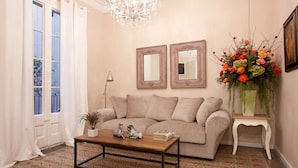 A living room with a beige sofa, a wooden coffee table, two framed mirrors, a chandelier, and a floral arrangement.