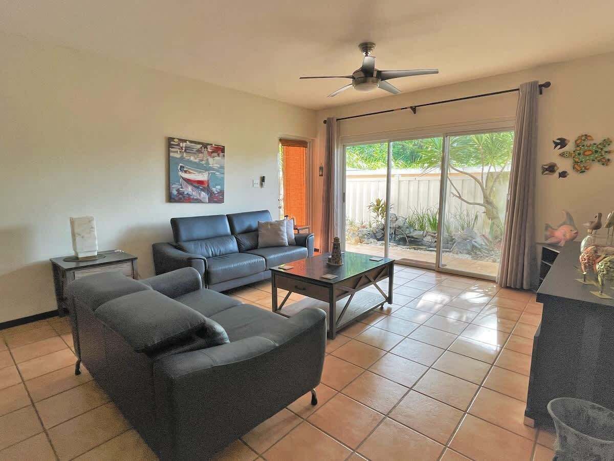 A living room with a sofa, coffee table, and a ceiling fan. There is a sliding glass door leading to a patio with plants.