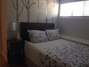Quarto com cama, mesa de cabeceira, abajur e decoração de árvores e pássaros na parede.