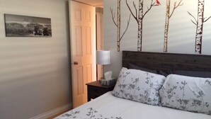 Une chambre à coucher comprenant un lit, une table de chevet, une lampe, une porte et un mur orné d’une fresque représentant un arbre et des oiseaux.