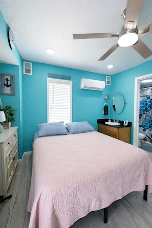 A bedroom with a bed, a ceiling fan, an air conditioning unit, a window with blinds, a sink, and a mirror.