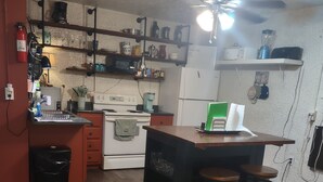 Una cocina con suelo de madera, estufa, refrigerador, ventilador de techo y una barra con taburetes.
