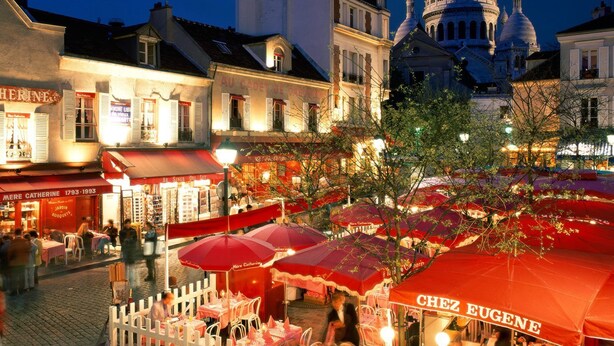A bustling street scene with outdoor seating and umbrellas, a prominent dome building in the background, and a variety of storefronts.