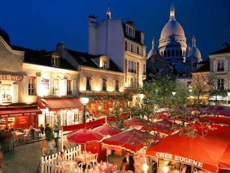 A bustling outdoor café area with tables and red umbrellas, surrounded by historic buildings and a prominent dome structure in the background.