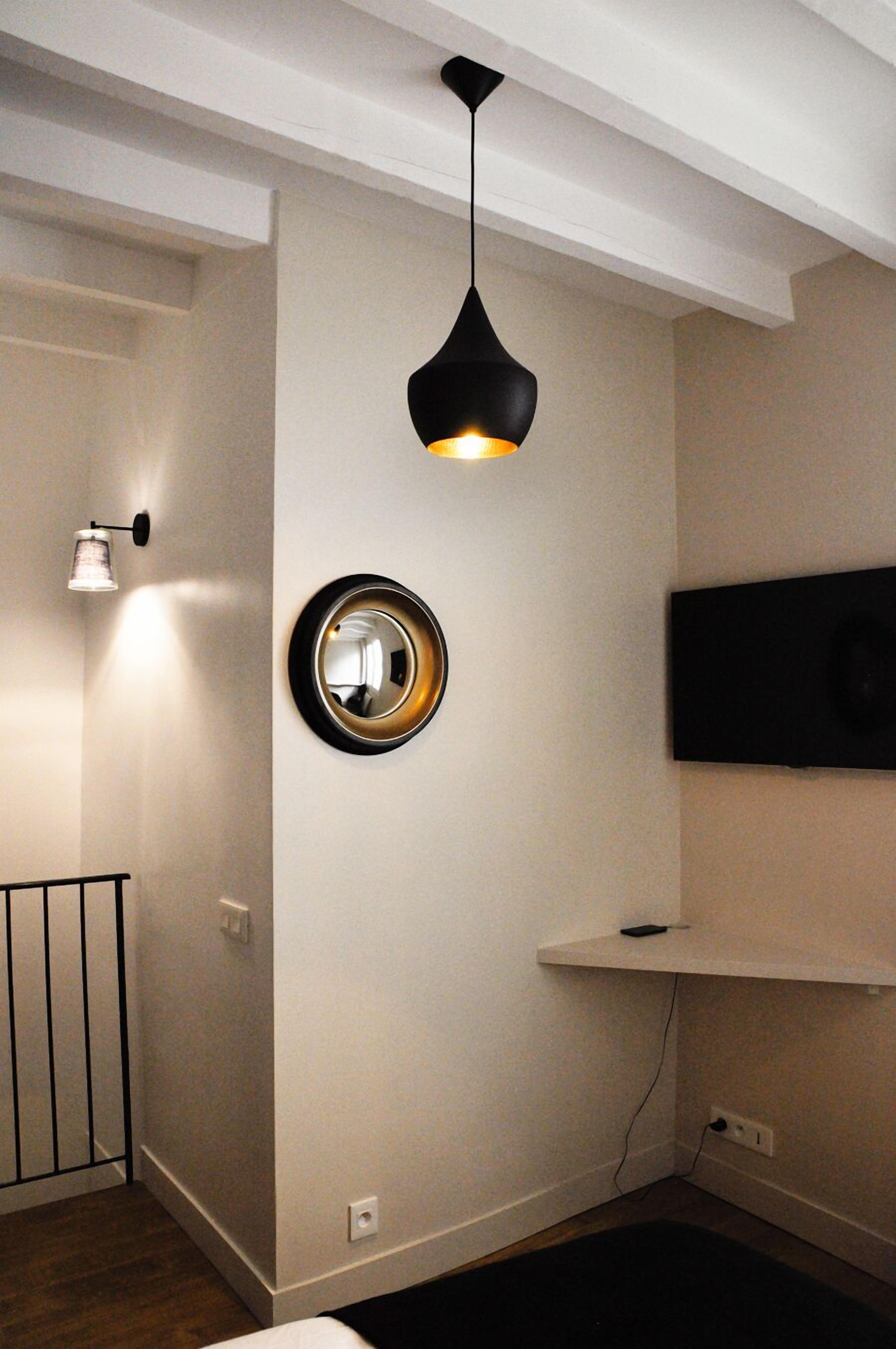 A room with a black pendant light, a round mirror, a flat-screen TV on a shelf, and a white wall with a door and a railing.