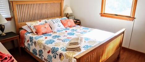 Una cama bien hecha con una colcha estampada y dos almohadas, una mesita de noche con una lámpara, y una ventana con persianas.