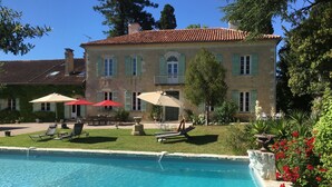 Une piscine avec des chaises longues et des parasols, un bâtiment en pierre aux volets verts, et un jardin parsemé de fleurs rouges.
