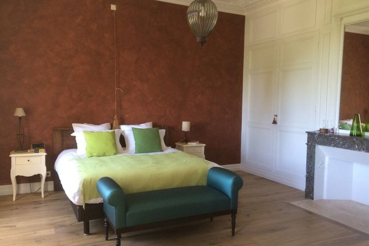 A bedroom with a bed, green and white pillows, a teal bench, a nightstand, a lamp, and a hanging light fixture.