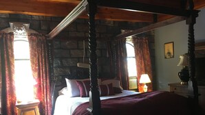 A four-poster canopy bed with a wooden headboard, a stone wall backdrop, and a nightstand with a lamp.