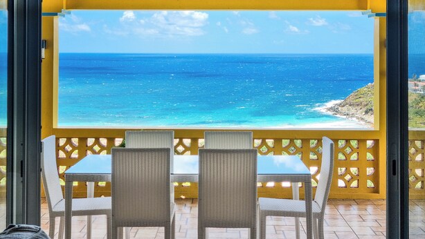 Ein Balkon mit Korbstühlen und einem Tisch, der auf einen Strand mit klarem, blauem Wasser und einer felsigen Küste blickt.