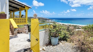 Ein gelbes Tor führt zu einem Balkon mit Blick auf einen Strand, kristallklares blaues Wasser und eine ferne Ansicht einer Stadt.
