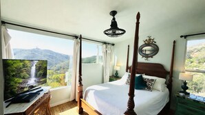 A four-poster bed with a view of a waterfall, a television on a wooden dresser, and a nightstand with a lamp.