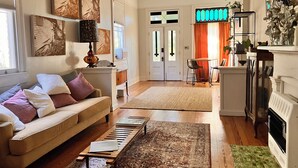 Un salon comprenant un canapé, une table basse et une cheminée. On y trouve également un coin repas avec une table et des chaises. La pièce est dotée de parquets et d’un ventilateur de plafond.