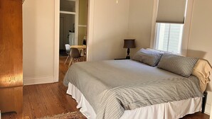 Une chambre avec un grand lit, une table de chevet, une lampe, une fenêtre avec des stores et un plancher en bois.