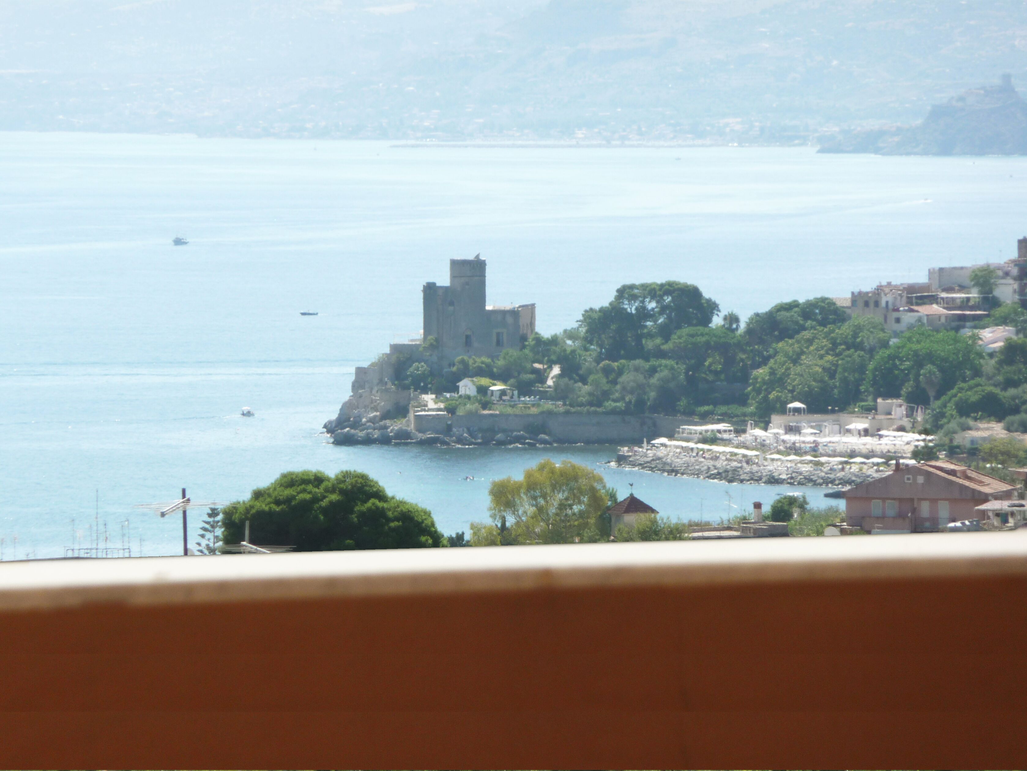 Una cittadina costiera con una struttura imponente simile a un castello, situata su uno sperone roccioso, circondata da vegetazione e edifici.