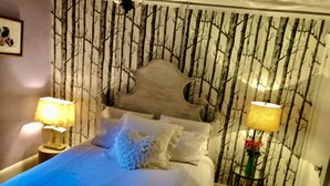 Un dormitorio con cama, dos lámparas de noche, una pared con estampado de árbol y una mesita con un jarrón.