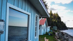 Rannalla sijaitseva mökki, jossa on suuri ikkuna, josta avautuu näkymä merelle, pieni terassi, jossa on tuoleja, sekä rantaviivan varrella oleva puu.