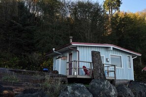 En lille, etetages hytte med rødt tag og hvide vægge, beliggende på en klippefyldt kyst med skov i baggrunden.