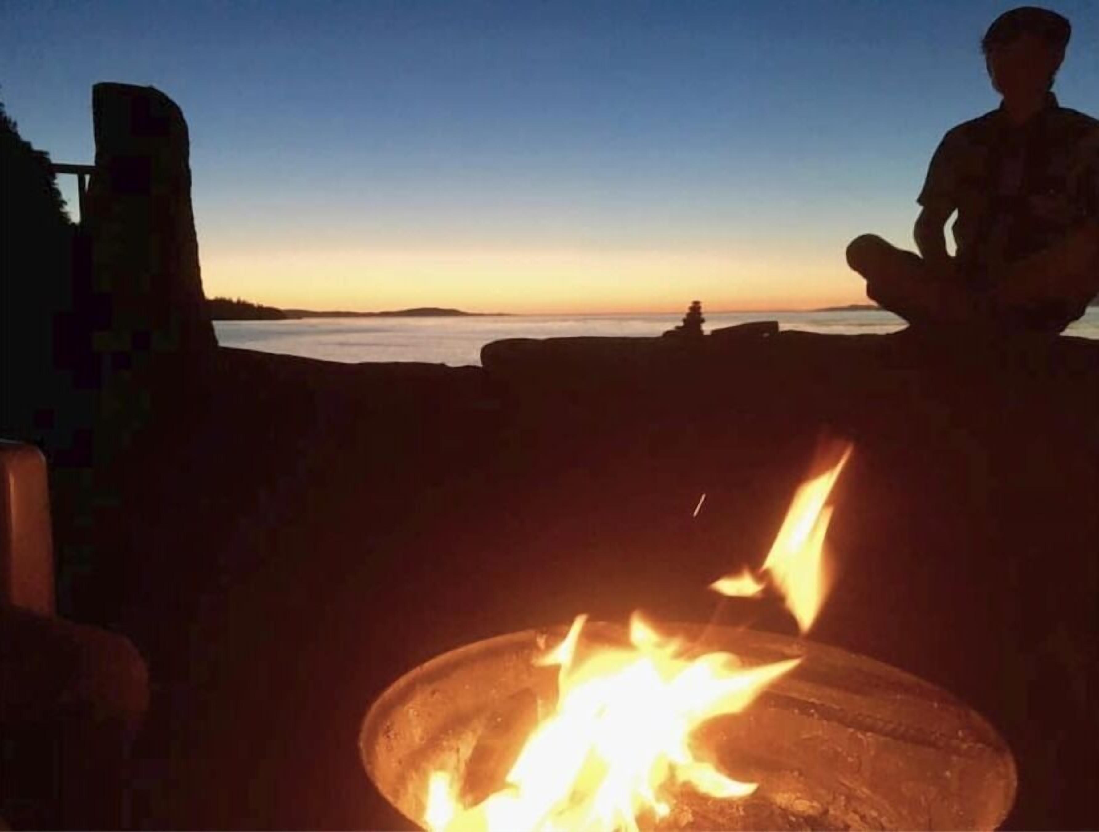 A campfire with a person sitting nearby, a body of water in the background, and a structure in the distance.