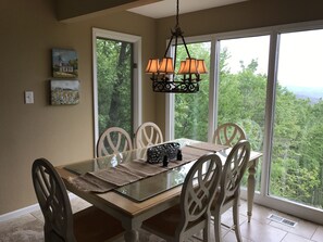 A dining area with a glass-top table, white chairs, a chandelier, and large windows offering a view of the forest.